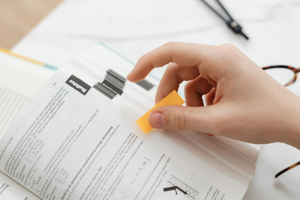 Student marking a textbook with a sticky note while studying for exams.