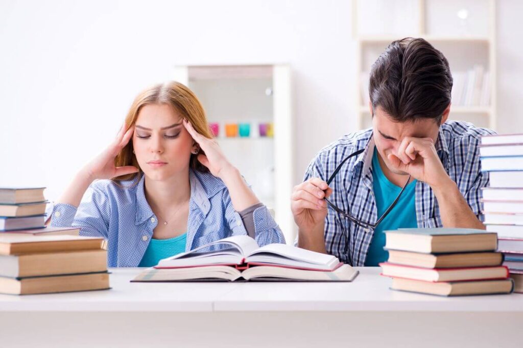 Students feeling stressed and tired while studying for exams surrounded by textbooks.