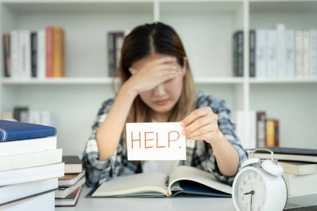 Stressed student studying for exams holding a sign that says help, surrounded by books and a clock.
